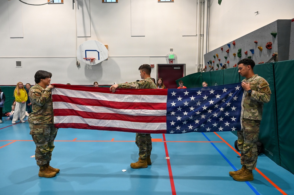 Three military personnel in OCP (Operational Camouflage Pattern) uniforms are performing a flag folding ceremony with an American flag inside a school gymnasium. Students can be seen seated along the walls observing the demonstration. The setting appears to be an educational outreach or Veterans Day-type event at a school facility.