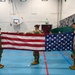 Three military personnel in OCP (Operational Camouflage Pattern) uniforms are performing a flag folding ceremony with an American flag inside a school gymnasium. Students can be seen seated along the walls observing the demonstration. The setting appears to be an educational outreach or Veterans Day-type event at a school facility.