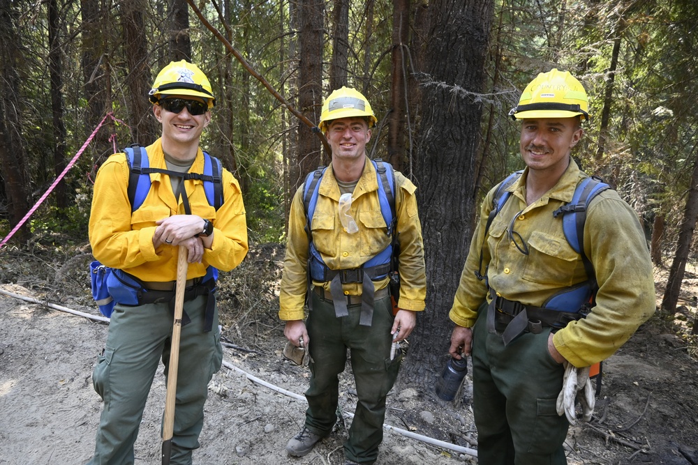 Three wildland firefighters are posing together in a forested area, wearing standard Nomex fire-resistant yellow shirts and green pants with yellow hard hats. Their gear shows varying degrees of dirt and soot, indicating active field work. They appear to be taking a break and are smiling at the camera.