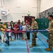 Three military personnel in Army Combat Uniforms (ACU) are demonstrating the proper folding of an American flag to a group of school-age children in a gymnasium. The demonstration appears to be an educational outreach event, with children watching attentively while holding small American flags. The setting is a school or community recreation facility equipped with a basketball hoop and a climbing wall.