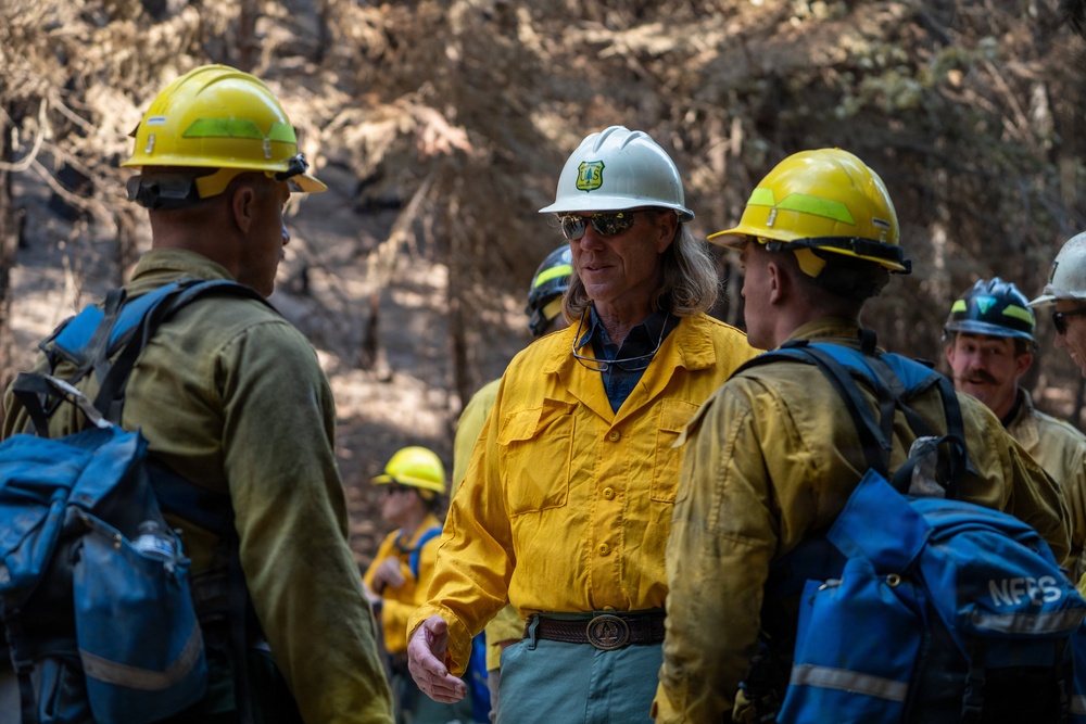 A group of wildland firefighters are gathered in a burned forest area, engaged in what appears to be a briefing or debriefing. The central figure wears a white hard hat with a US Forest Service shield emblem and a yellow Nomex fire-resistant shirt, indicating a supervisory or leadership role. Other crew members wearing yellow Nomex shirts and yellow hard hats carry blue backpacks labeled 'NFFS'.