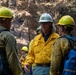 A group of wildland firefighters are gathered in a burned forest area, engaged in what appears to be a briefing or debriefing. The central figure wears a white hard hat with a US Forest Service shield emblem and a yellow Nomex fire-resistant shirt, indicating a supervisory or leadership role. Other crew members wearing yellow Nomex shirts and yellow hard hats carry blue backpacks labeled 'NFFS'.