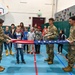Three military personnel in OCP (Operational Camouflage Pattern) uniforms are demonstrating the proper folding of an American flag to a group of school-age children in a gymnasium. The demonstration appears to be an educational outreach event, likely tied to a patriotic or Veterans Day program. The children are attentively watching as the soldiers carefully fold the flag in the traditional triangular method.