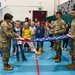 Three military personnel in OCP (Operational Camouflage Pattern) uniforms are demonstrating the proper folding of an American flag to a group of elementary school-aged children inside a gymnasium. The demonstration appears to be an educational event, likely tied to a patriotic or Veterans Day program. The setting is a school gym equipped with a basketball hoop, climbing wall, and padded walls.