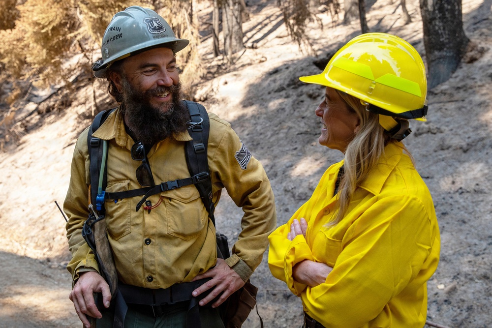 Two wildland firefighters are engaged in a friendly conversation in a fire-affected forest area. The man on the left wears a white hard hat with partial text reading 'TWAIN' and 'CREW,' along with a yellow Nomex shirt and a backpack with radio equipment. The woman on the right wears a bright yellow hard hat and matching yellow Nomex shirt with her arms crossed.