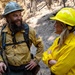 Two wildland firefighters are engaged in a friendly conversation in a fire-affected forest area. The man on the left wears a white hard hat with partial text reading 'TWAIN' and 'CREW,' along with a yellow Nomex shirt and a backpack with radio equipment. The woman on the right wears a bright yellow hard hat and matching yellow Nomex shirt with her arms crossed.