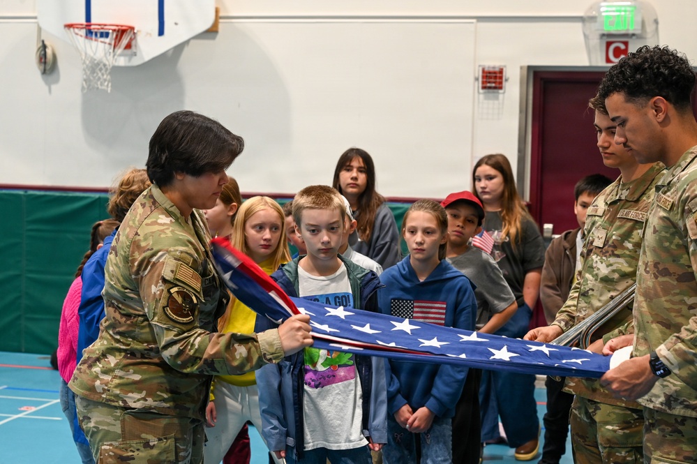 Two U.S. Air Force service members in OCP (Operational Camouflage Pattern) uniforms are demonstrating the proper folding of an American flag to a group of school-aged children in what appears to be a school gymnasium. The educational outreach event shows the military personnel actively engaging with students, with one service member holding the partially folded flag while the other assists. Several children are watching attentively, with one girl wearing a hoodie featuring an American flag patch.