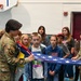 Two U.S. Air Force service members in OCP (Operational Camouflage Pattern) uniforms are demonstrating the proper folding of an American flag to a group of school-aged children in what appears to be a school gymnasium. The educational outreach event shows the military personnel actively engaging with students, with one service member holding the partially folded flag while the other assists. Several children are watching attentively, with one girl wearing a hoodie featuring an American flag patch.
