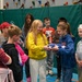 A group of school-age children are gathered in what appears to be a school gymnasium with a climbing wall in the background. Two girls are the focal point — one in a yellow hoodie and one in a blue hoodie — jointly holding a folded American flag in the traditional triangular ceremonial fold. The scene suggests a patriotic educational activity, possibly related to Veterans Day, Memorial Day, or a civic lesson.