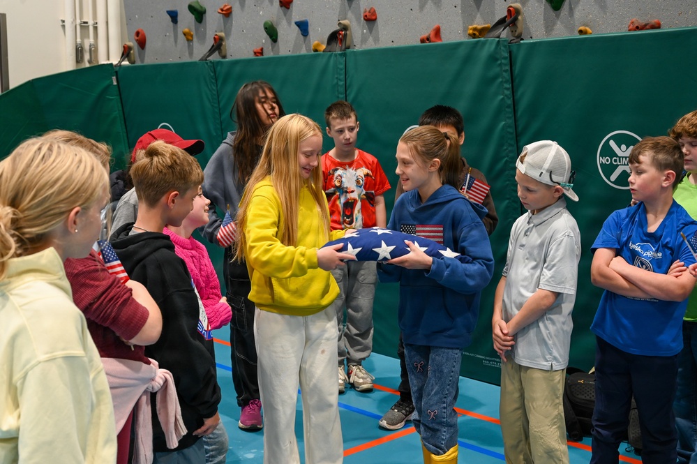 A group of elementary school-aged children are gathered in a school gymnasium with a climbing wall in the background. Two children at the center are holding a folded American flag together, while surrounding students observe, some holding small American flags. The scene appears to be part of a patriotic educational activity or ceremony.