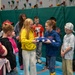 A group of elementary school-aged children are gathered in a school gymnasium with a climbing wall in the background. Two children at the center are holding a folded American flag together, while surrounding students observe, some holding small American flags. The scene appears to be part of a patriotic educational activity or ceremony.