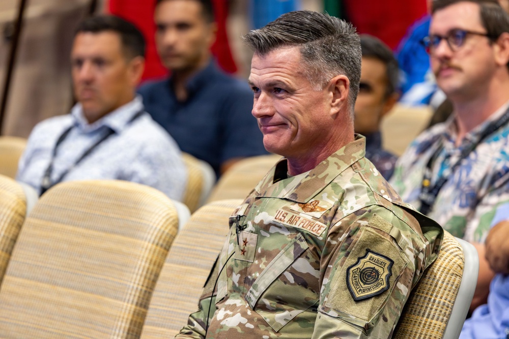 A U.S. Air Force officer in OCP (Operational Camouflage Pattern) uniform is seated in an auditorium or meeting room, smiling slightly. He displays two stars on his chest indicating the rank of Major General, along with a 'Graduate' patch on his sleeve. Several civilians and possibly other military personnel are seated behind him in a blurred background.