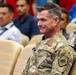 A U.S. Air Force officer in OCP (Operational Camouflage Pattern) uniform is seated in an auditorium or meeting room, smiling slightly. He displays two stars on his chest indicating the rank of Major General, along with a 'Graduate' patch on his sleeve. Several civilians and possibly other military personnel are seated behind him in a blurred background.