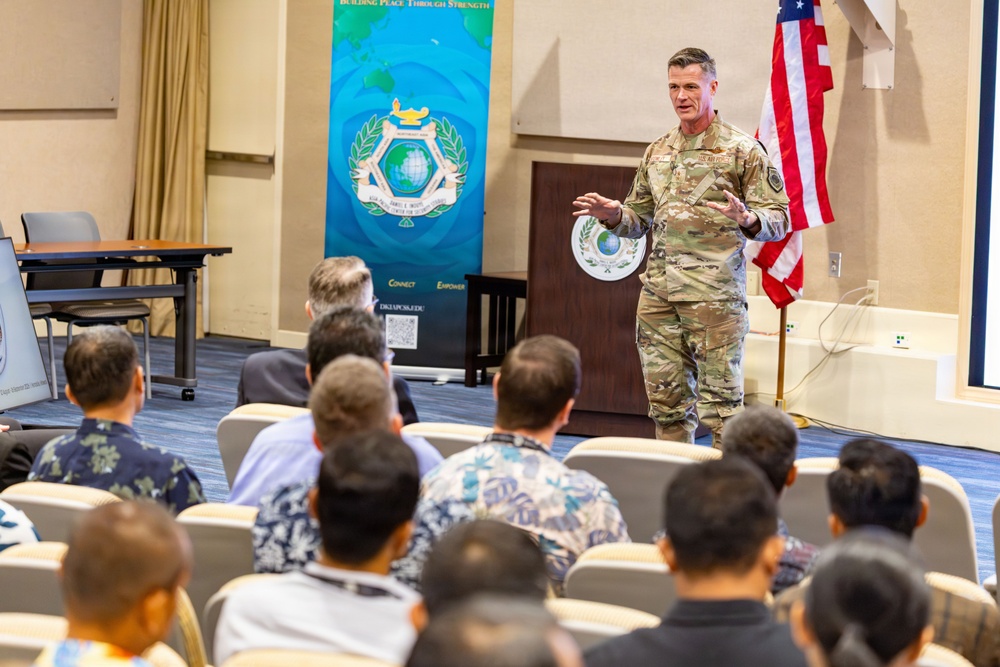 A U.S. military officer in camouflage uniform is addressing an audience in a conference or briefing room setting. The speaker stands near a podium beside a U.S. flag and a banner reading 'Building Peace Through Strength' with the text 'Connect' and 'Empower' visible. The audience, seated in rows of chairs, appears to include both military and civilian attendees, several wearing Hawaiian-style shirts.