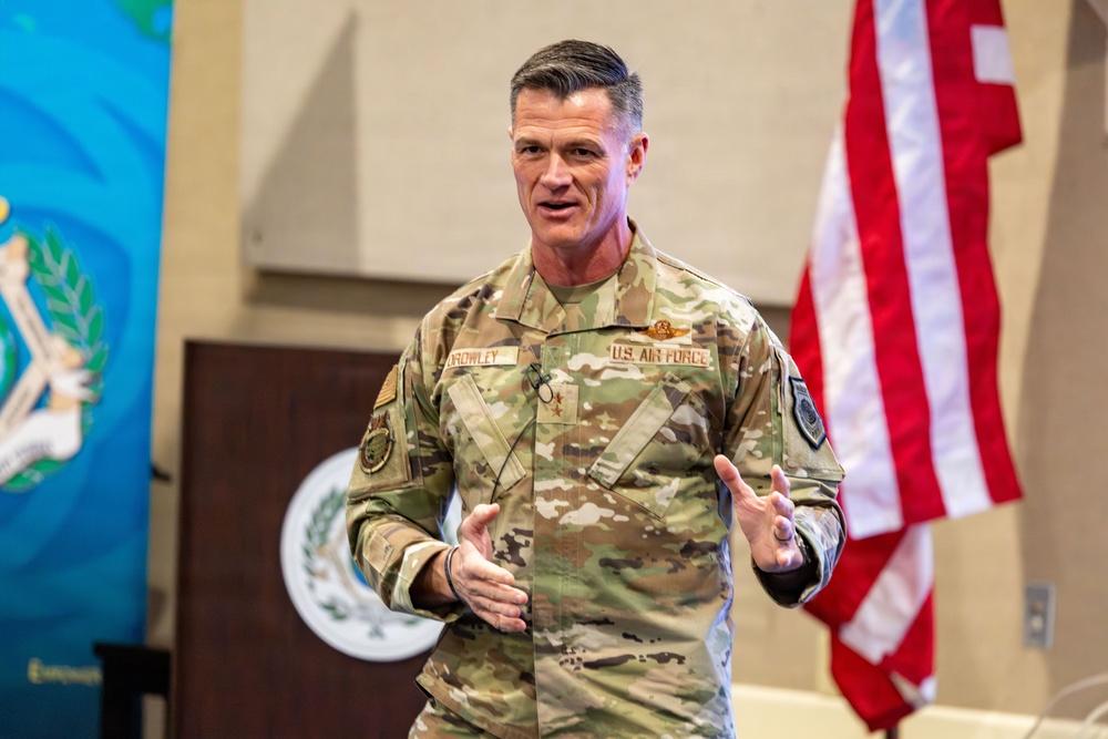 A U.S. Air Force officer wearing OCP (Operational Camouflage Pattern) uniform with the name tape 'CROWLEY' and a one-star general rank insignia is speaking at what appears to be an official military briefing or presentation. He is gesturing with both hands, suggesting active engagement with an audience. An American flag and what appears to be an official seal or emblem are visible in the background.
