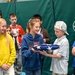 A group of school-age children are gathered indoors, likely in a gymnasium or recreation center, participating in what appears to be a patriotic ceremony or educational activity. Two children in the center are handling a folded American flag, with one child in a blue hoodie featuring an American flag patch presenting or receiving it from a boy in a white cap. The other children are watching attentively, suggesting a structured, meaningful event.
