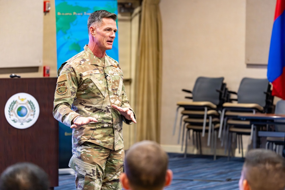 A U.S. Army officer in OCP (Operational Camouflage Pattern) uniform is delivering a presentation or briefing to an audience in what appears to be a conference or briefing room. He is wearing a lapel microphone and gesturing with his hands while speaking. The setting includes institutional signage, a podium with an emblem, and what appears to be a Mongolian flag in the background.