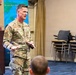 A U.S. Army officer in OCP (Operational Camouflage Pattern) uniform is delivering a presentation or briefing to an audience in what appears to be a conference or briefing room. He is wearing a lapel microphone and gesturing with his hands while speaking. The setting includes institutional signage, a podium with an emblem, and what appears to be a Mongolian flag in the background.