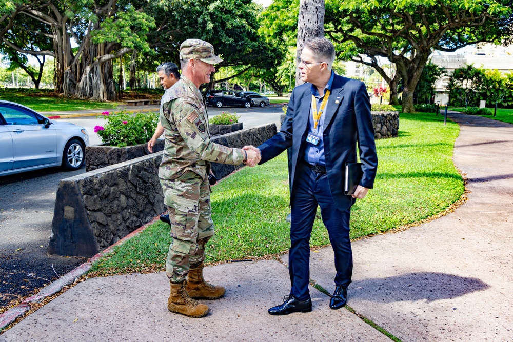 A U.S. Army soldier in OCP (Operational Camouflage Pattern) uniform is shaking hands with a civilian man wearing a dark suit and a lanyard/badge on a paved walkway. The setting appears to be a military installation, likely in Hawaii based on the tropical vegetation including large banyan trees and lush greenery. The interaction appears to be a formal greeting or welcome.