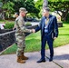 A U.S. Army soldier in OCP (Operational Camouflage Pattern) uniform is shaking hands with a civilian man wearing a dark suit and a lanyard/badge on a paved walkway. The setting appears to be a military installation, likely in Hawaii based on the tropical vegetation including large banyan trees and lush greenery. The interaction appears to be a formal greeting or welcome.