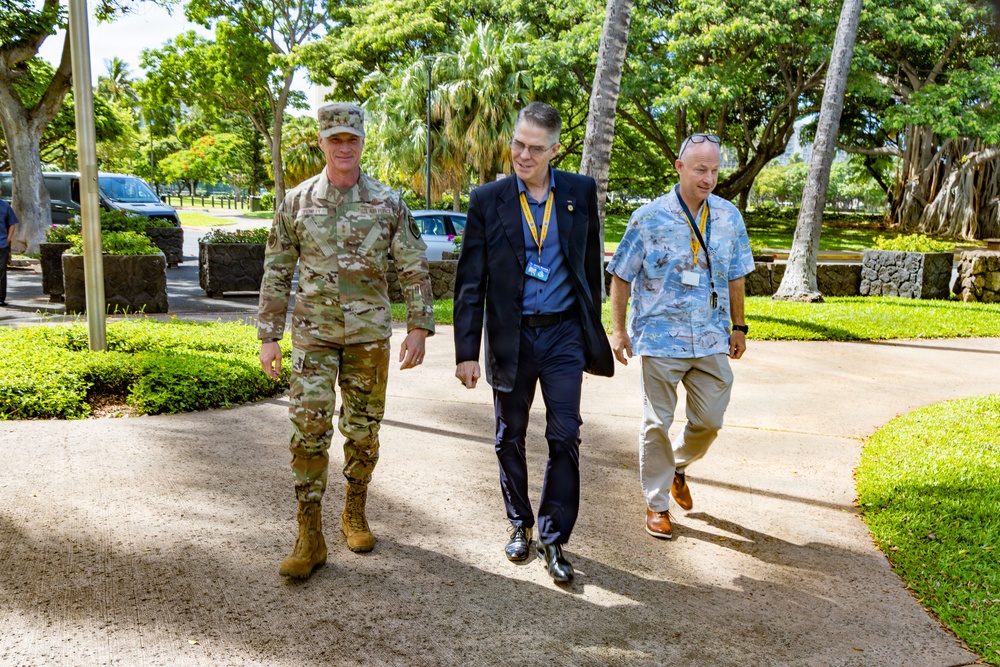 Three men are walking together along a paved pathway in a lush, tropical outdoor setting. One man is wearing military camouflage uniform with combat boots, while the other two are in civilian attire with visible lanyards and badges. The setting appears to be a military installation or government facility based on the manicured grounds and flagpole visible in the background.