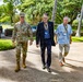 Three men are walking together along a paved pathway in a lush, tropical outdoor setting. One man is wearing military camouflage uniform with combat boots, while the other two are in civilian attire with visible lanyards and badges. The setting appears to be a military installation or government facility based on the manicured grounds and flagpole visible in the background.