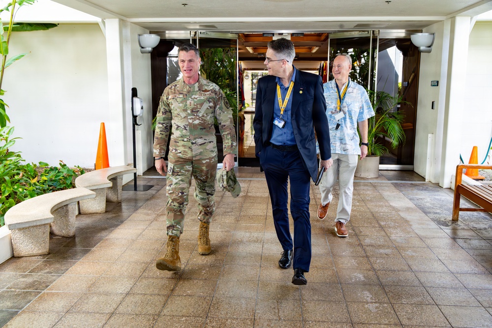 Three individuals are walking together through what appears to be a covered outdoor entrance or portico of a military or government facility. One person is wearing U.S. military camouflage (OCP) uniform with tan boots, while the other two are in civilian attire wearing lanyards with identification badges. The setting appears to be in a tropical location based on the vegetation and architectural style.