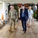 Three individuals are walking together through what appears to be a covered outdoor entrance or portico of a military or government facility. One person is wearing U.S. military camouflage (OCP) uniform with tan boots, while the other two are in civilian attire wearing lanyards with identification badges. The setting appears to be in a tropical location based on the vegetation and architectural style.