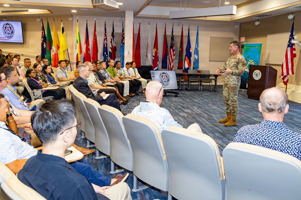 A military officer in camouflage uniform is addressing a diverse audience of civilians and military personnel in a formal conference room. The setting appears to be a multinational security cooperation conference, evidenced by the numerous national flags displayed along the back wall and a presentation screen showing a conference logo. The audience is seated in rows of chairs and appears engaged with the speaker.