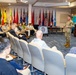 A military officer in camouflage uniform is addressing a diverse audience of civilians and military personnel in a formal conference room. The setting appears to be a multinational security cooperation conference, evidenced by the numerous national flags displayed along the back wall and a presentation screen showing a conference logo. The audience is seated in rows of chairs and appears engaged with the speaker.