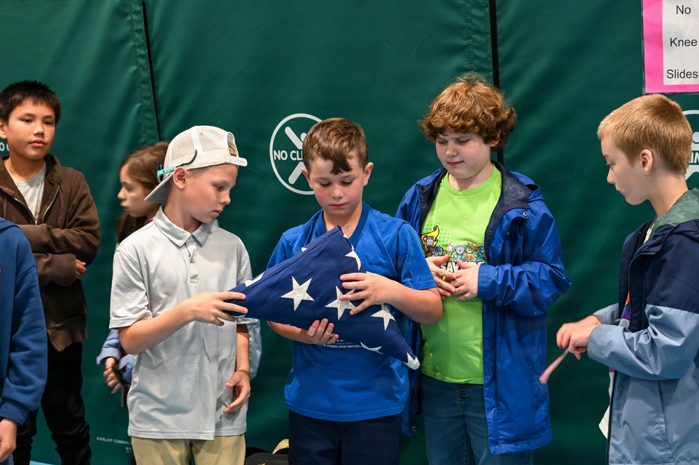A group of elementary school-aged children are gathered indoors, with two boys at the center carefully holding a folded American flag. The setting appears to be a gymnasium or recreational facility, suggested by the padded green walls and posted rules sign. This appears to be an educational or ceremonial activity involving proper flag handling.