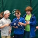 A group of elementary school-aged children are gathered indoors, with two boys at the center carefully holding a folded American flag. The setting appears to be a gymnasium or recreational facility, suggested by the padded green walls and posted rules sign. This appears to be an educational or ceremonial activity involving proper flag handling.
