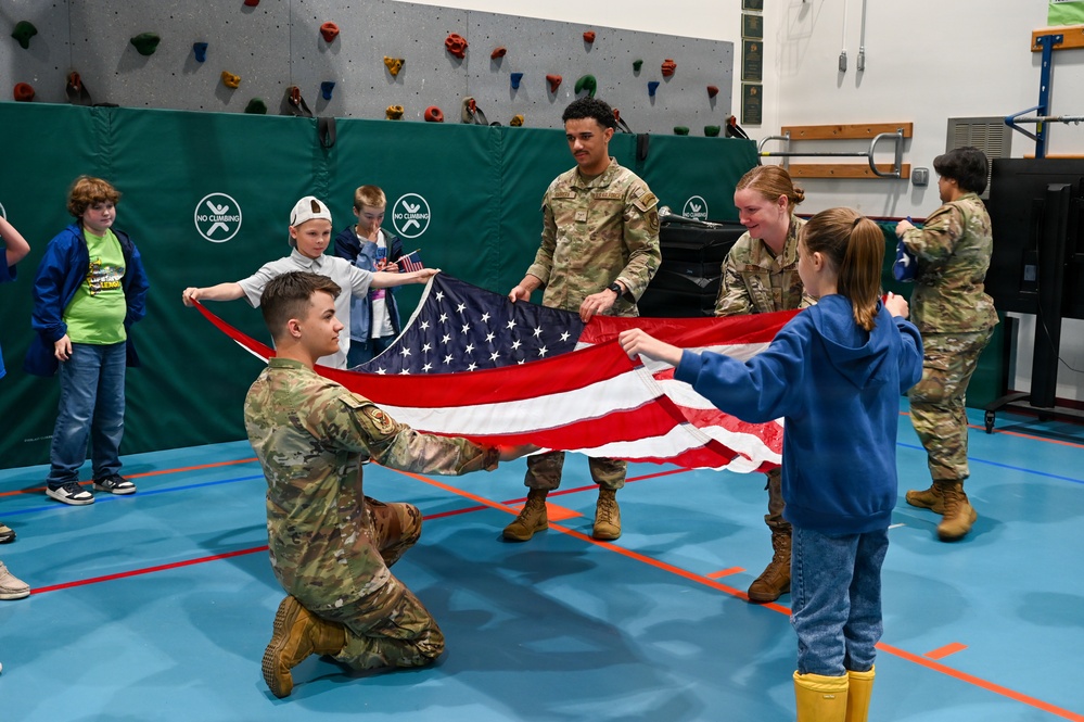 Military personnel in camouflage uniforms are teaching children how to properly fold an American flag inside what appears to be a school gymnasium. The activity involves multiple soldiers working alongside several children, demonstrating flag folding protocol in an educational setting. A rock climbing wall with safety padding is visible in the background.