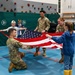 Military personnel in camouflage uniforms are teaching children how to properly fold an American flag inside what appears to be a school gymnasium. The activity involves multiple soldiers working alongside several children, demonstrating flag folding protocol in an educational setting. A rock climbing wall with safety padding is visible in the background.