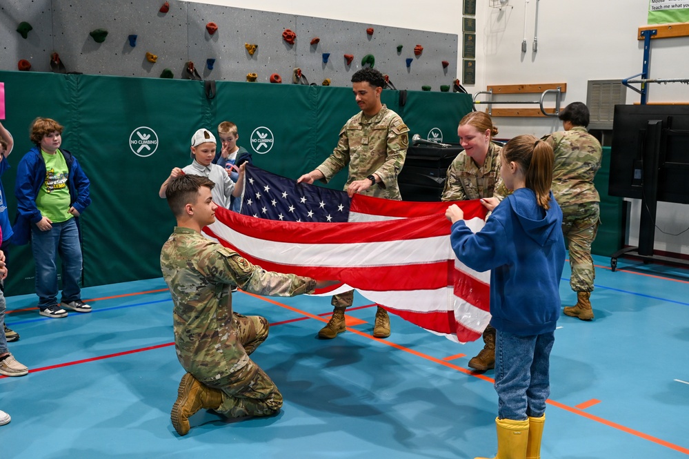 Military personnel in camouflage uniforms are teaching children how to fold an American flag inside what appears to be a school gymnasium with a rock climbing wall. Three soldiers are actively demonstrating the flag folding process with at least one child participating directly. The setting suggests an educational outreach or community engagement event.