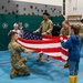 Military personnel in camouflage uniforms are teaching children how to fold an American flag inside what appears to be a school gymnasium with a rock climbing wall. Three soldiers are actively demonstrating the flag folding process with at least one child participating directly. The setting suggests an educational outreach or community engagement event.
