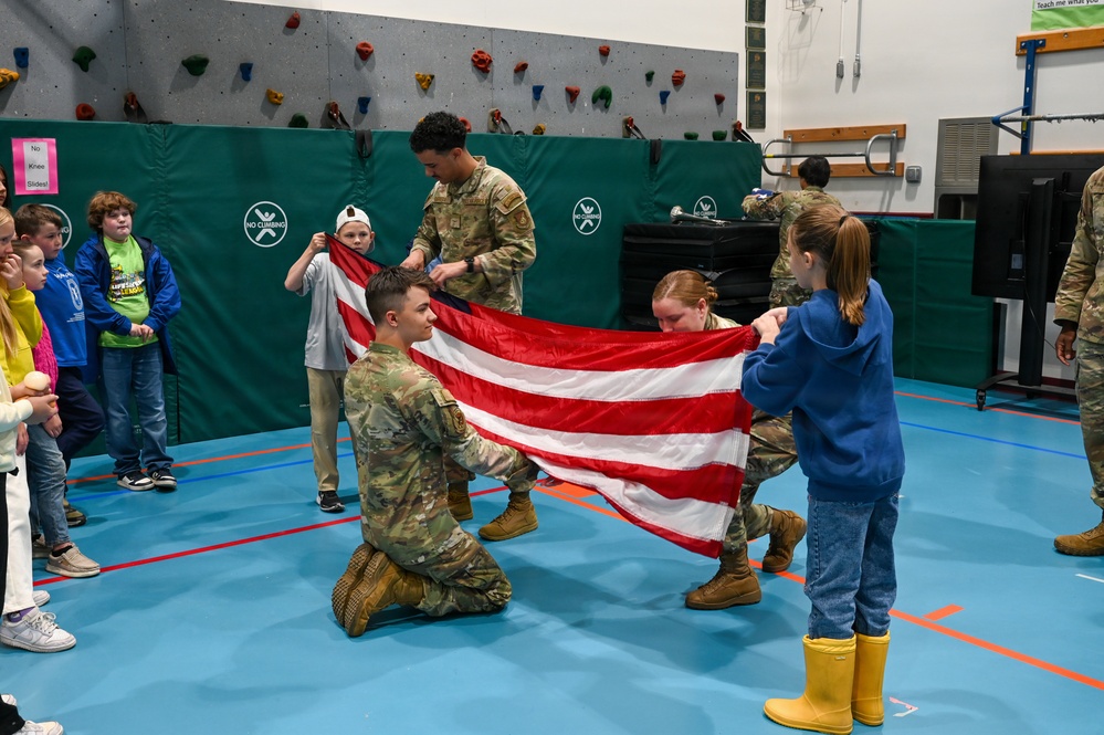 Military personnel in camouflage uniforms are demonstrating flag folding to children in what appears to be a school gymnasium. Two or three soldiers are working together with at least one child to fold an American flag, while other children watch from the sides. This appears to be an educational outreach or Veterans Day-type event at an elementary school.
