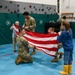 Military personnel in camouflage uniforms are demonstrating flag folding to children in what appears to be a school gymnasium. Two or three soldiers are working together with at least one child to fold an American flag, while other children watch from the sides. This appears to be an educational outreach or Veterans Day-type event at an elementary school.