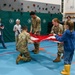 Two military personnel in camouflage uniforms are demonstrating the proper folding of an American flag to elementary school children in a gymnasium. A child in a blue sweatshirt and yellow boots is actively participating in the flag folding alongside the soldiers. Other students observe from the sides while one child holds a small American flag.