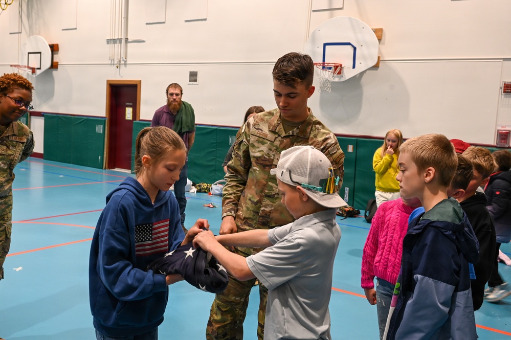 A military service member in camouflage uniform is teaching children how to fold an American flag in what appears to be a school gymnasium. Two children are actively handling the flag while others observe. This appears to be an educational outreach or patriotic demonstration event.