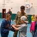 A military service member in camouflage uniform is teaching children how to fold an American flag in what appears to be a school gymnasium. Two children are actively handling the flag while others observe. This appears to be an educational outreach or patriotic demonstration event.