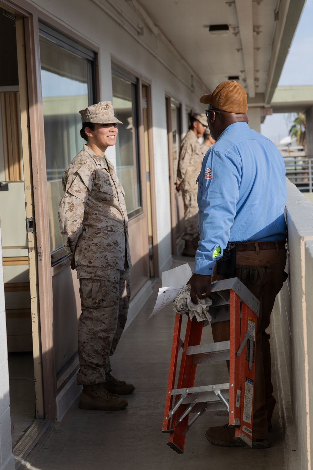 A U.S. Marine in desert MARPAT camouflage uniform is conversing with a civilian maintenance or contractor worker on an outdoor corridor of a multi-story building. The civilian is holding an orange step ladder and wearing a blue work shirt with a company patch on the sleeve. Additional Marines in uniform are visible in the background of the same corridor.