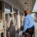 A U.S. Marine in desert MARPAT camouflage uniform is conversing with a civilian maintenance or contractor worker on an outdoor corridor of a multi-story building. The civilian is holding an orange step ladder and wearing a blue work shirt with a company patch on the sleeve. Additional Marines in uniform are visible in the background of the same corridor.