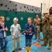A group of children and military personnel in camouflage uniforms are gathered in a school gymnasium with a rock climbing wall in the background. A young girl wearing a blue American flag hoodie and yellow rain boots is holding a folded American flag, while military members and other children applaud around her. The scene appears to be a ceremonial presentation of a flag to the child.
