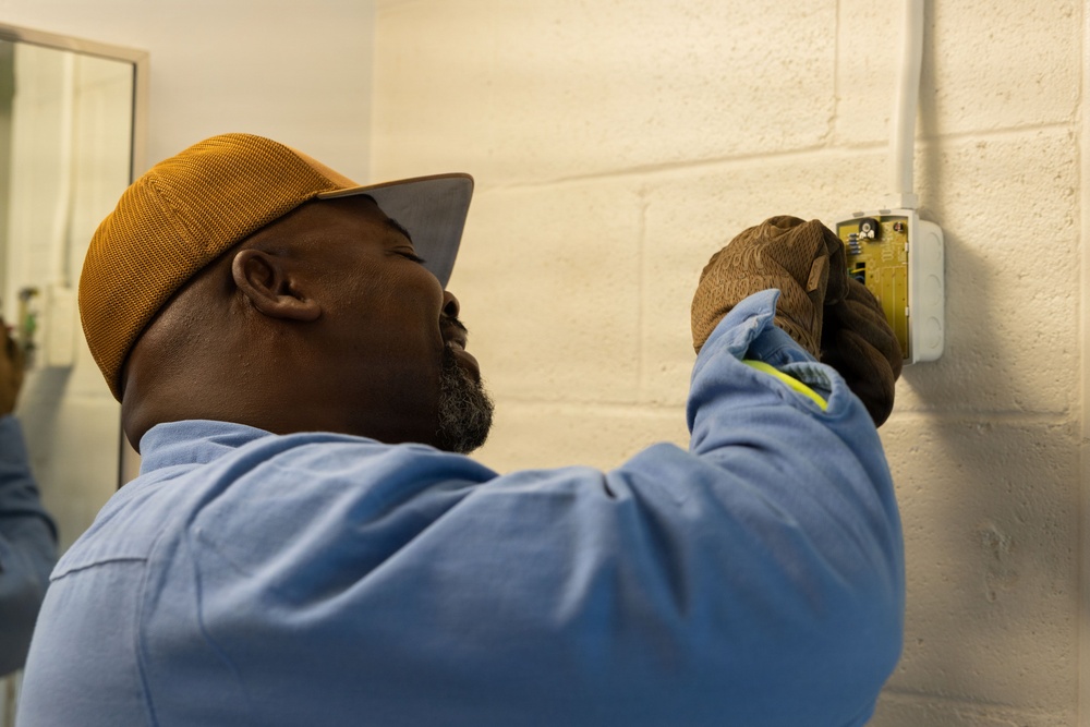 A man wearing a gold/mustard baseball cap and blue work shirt is installing or servicing an electrical component on a painted cinder block wall. He is wearing work gloves and appears to be carefully handling a wall-mounted electrical box or device. The image is taken from a close side-angle perspective, emphasizing the focused nature of the task.