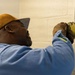 A man wearing a gold/mustard baseball cap and blue work shirt is installing or servicing an electrical component on a painted cinder block wall. He is wearing work gloves and appears to be carefully handling a wall-mounted electrical box or device. The image is taken from a close side-angle perspective, emphasizing the focused nature of the task.