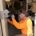 A middle-aged man wearing an orange long-sleeve shirt and tan cargo pants is crouched down working on a door lock or handle mechanism. He appears to be a maintenance worker or locksmith, installing or repairing hardware on a door. The setting appears to be a military barracks or dormitory-style facility, with bunk beds and tactical gear visible in the background.