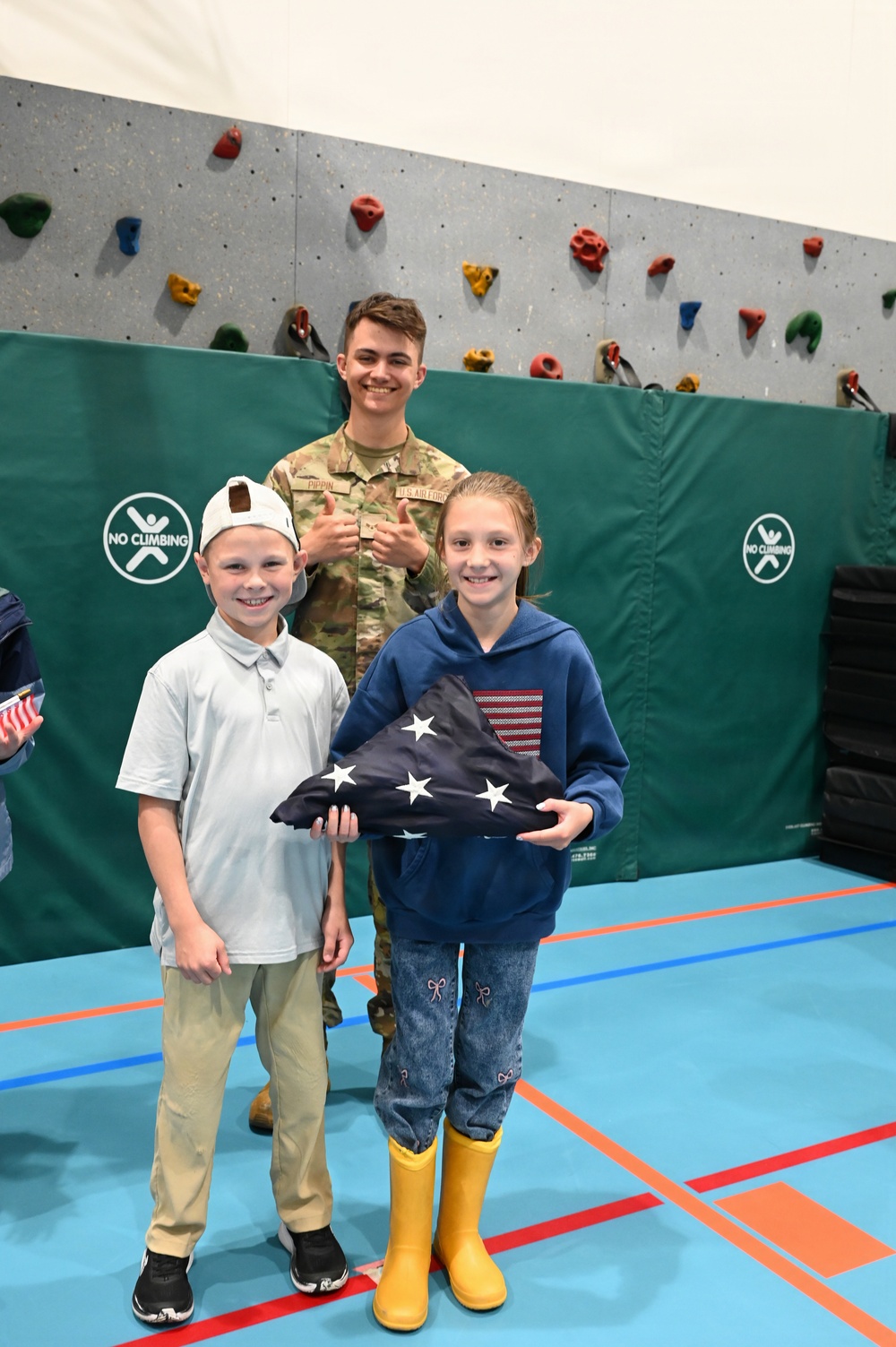A U.S. Air Force service member in camouflage uniform poses with two children in what appears to be a school or recreational gymnasium. The girl is holding a ceremonially folded American flag, suggesting a patriotic or Veterans Day-related event. The service member's name tape reads 'PIPPIN' and the branch tape reads 'U.S. AIR FORCE'.