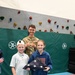 A U.S. Air Force service member in camouflage uniform poses with two children in what appears to be a school or recreational gymnasium. The girl is holding a ceremonially folded American flag, suggesting a patriotic or Veterans Day-related event. The service member's name tape reads 'PIPPIN' and the branch tape reads 'U.S. AIR FORCE'.