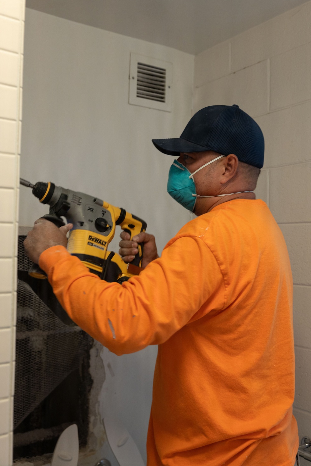 A worker wearing an orange long-sleeve shirt, dark blue baseball cap, and teal respiratory mask is using a DeWalt rotary hammer drill on a white-painted concrete block wall. The setting appears to be an interior room undergoing renovation or construction work. Dust and debris are visible at the base of the wall where drilling is occurring.